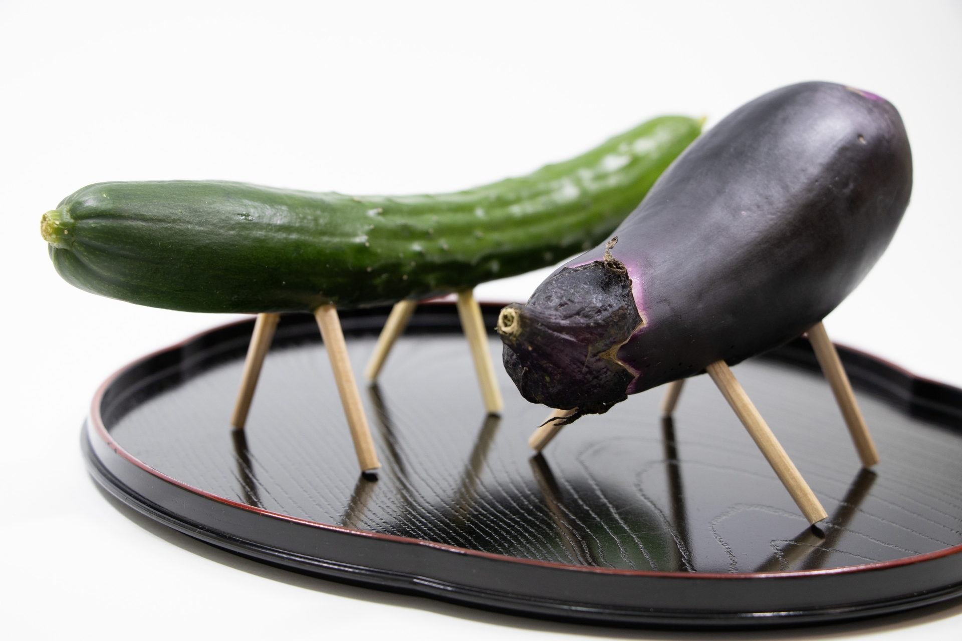 A cucumber horse and eggplant cow with wooden legs displayed on a black tray for Obon