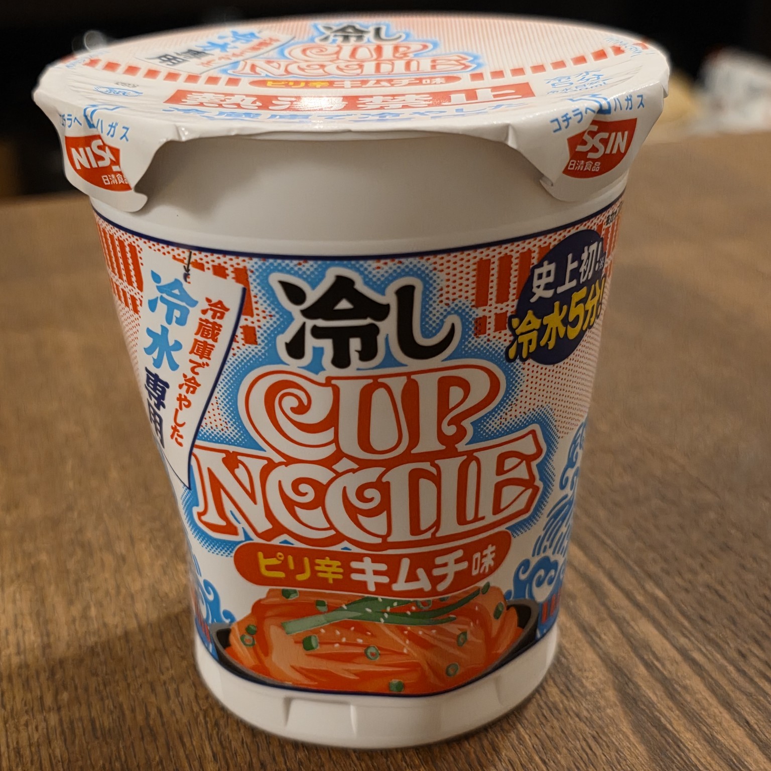 Front view of Nissin Hiyashi Cup Noodle spicy kimchi flavor on a wooden table, showing the Japanese cold-water preparation branding.
